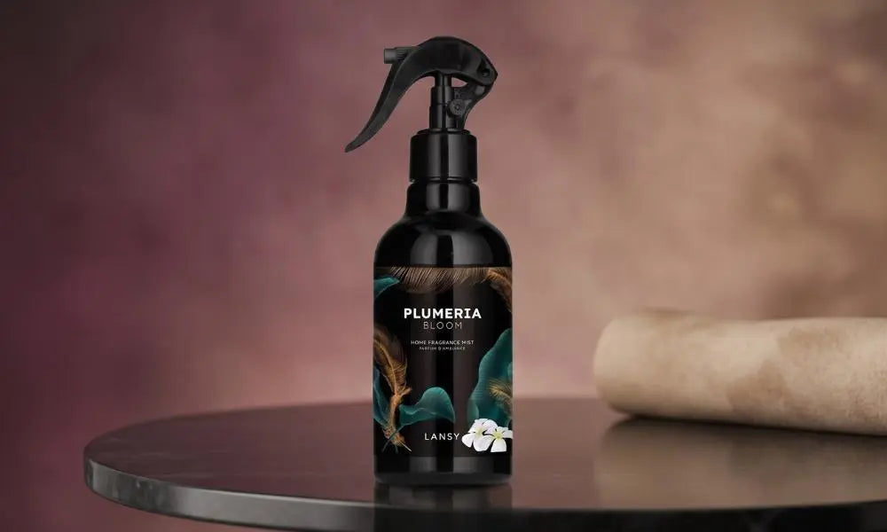 A centered black spray bottle of "PLUMERIA BLOOM" home fragrance mist on a dark, reflective surface, against a soft gradient background.