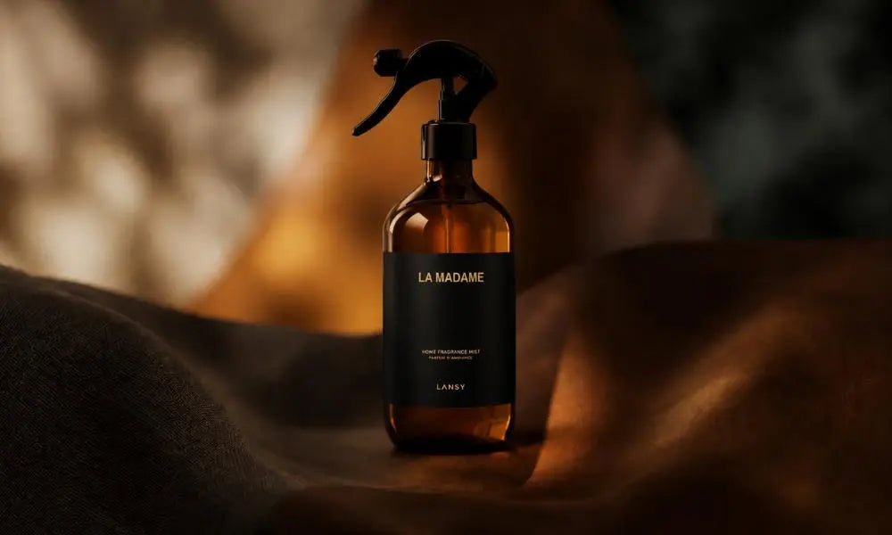 An amber spray bottle labeled 'LA MADAME' rests on a dark surface, surrounded by rich brown leather and textile folds under golden light.