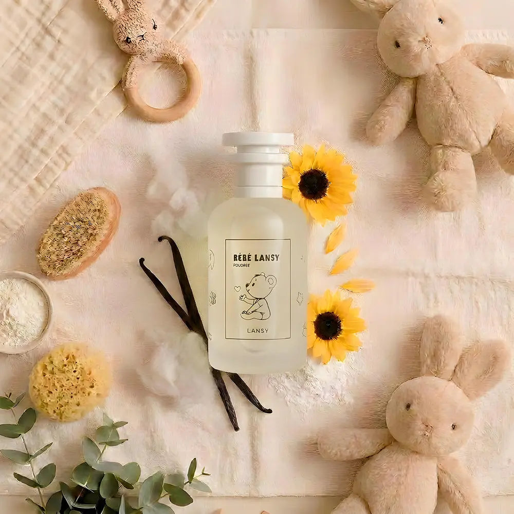 A frosted glass bottle labeled 'BÎBE LANSY' sits on a soft cream fabric surrounded by baby-themed props, including plush bunny toys, a wooden ring rattle, sponge brushes, vanilla beans, white powder, and small yellow flowers.