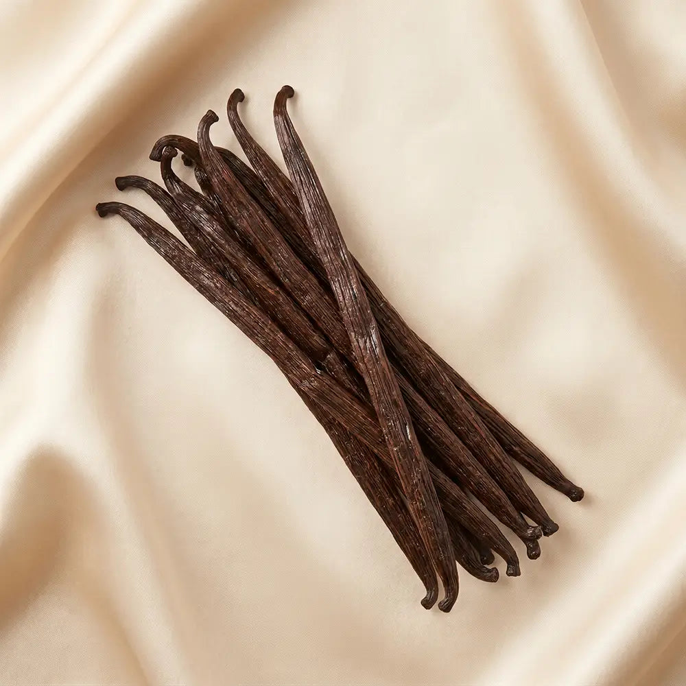 Dark brown vanilla beans arranged on draped cream satin fabric.