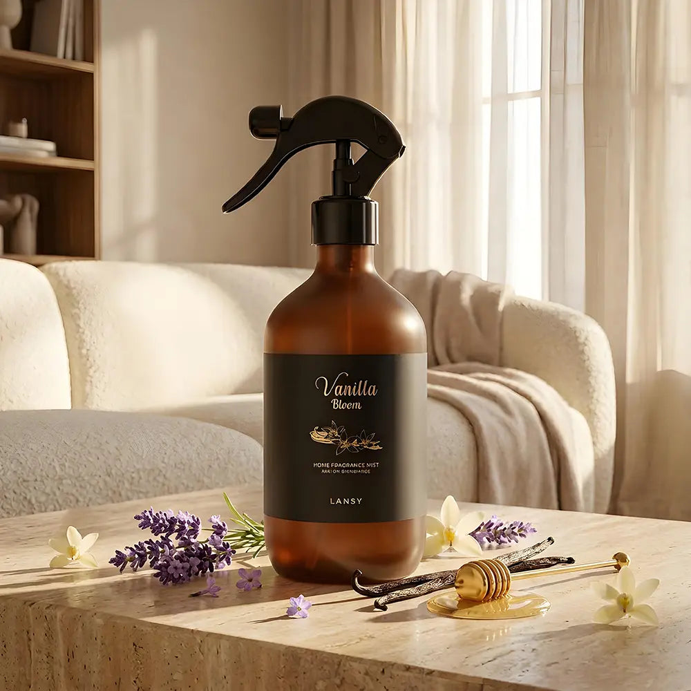 A bottle of Lansy Vanilla Bloom room fragrance mist sits on a wooden table in a cozy, sunlit living room setting. The amber glass bottle has a black spray nozzle and a black label with gold text.